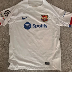 Vit FC Barcelona matchtröja Nike - Säljer en vit FC Barcelona matchtröja med blå detaljer och tryck. Tröjan har Spotify-logga på bröstet, Champions League-märke på ärmen och spelarnamn R. Araujo samt nummer 4 på ryggen. Tillverkad i funktionsmaterial, perfekt för fotboll eller träning.