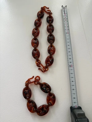 Bärnstensfärgat chunky armband och halsband  - Säljer ett chunky armband med stora ovala länkar i bärnstensfärgad plast med mörkbruna inslag. Armbandet har en cool retrovibe och länkarna är blanka och genomskinliga. Perfekt accessoar för att lyfta din outfit.