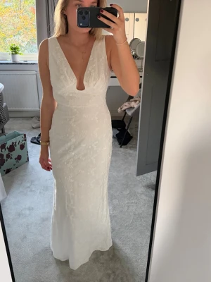 Adoore klänning  - Elegant vit långklänning med djup v-ringning och liten cut-out framtill. Klänningen har fina spetsdetaljer över hela tyget och är ärmlös. Perfekt för speciella tillfällen när du vill känna dig extra fin.