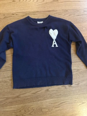 Ami Paris tröja - Snygg mörkblå stickad tröja från Ami Alexandre Mattiussi med stor vit hjärtlogga och bokstaven A på bröstet. Klassisk rund halsringning och långa ärmar. Perfekt för dig som gillar stilrena plagg med en twist. Dock är det ett fixat hål på baksidan.