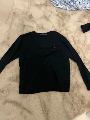 Svart tröja från Polo Ralph Lauren - Klassisk svart långärmad tröja från Polo Ralph Lauren. Tröjan har rund halsringning och en liten röd broderad logga på bröstet. Tillverkad i mjukt bomullsmaterial som känns skönt mot huden. Perfekt för en clean och stilren look.
