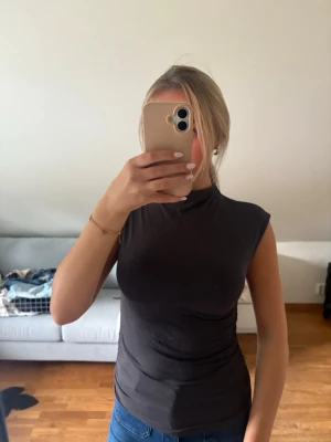 Mörkgrå ärmlös polotopp - Säljer en mörkgrå, tight polotopp utan ärmar. Toppen har en hög hals och är gjord i ett mjukt, stretchigt material som sitter snyggt mot kroppen. Perfekt att styla med jeans eller kjol för en clean look.