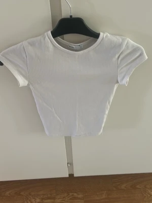 Vit ribbad croppad t-shirt - Säljer en enkel och stilren vit t-shirt med ribbad struktur och croppad passform. T-shirten har korta ärmar och rund halsringning, perfekt till jeans eller kjol för en clean look.