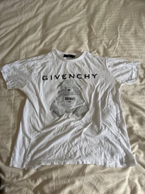 Vit Givenchy t-shirt med björntryck - Säljer en vit t-shirt från Givenchy med ett stort tryck av en björn och Givenchy-logga på bröstet. T-shirten har rund hals och korta ärmar. Perfekt för dig som gillar streetwear och vill ha något från ett lyxigt märke.