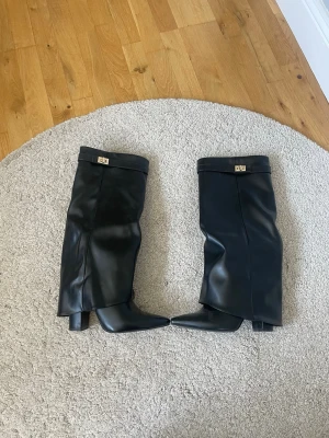 Svarta boots från Lululia  - Säljer ett par svarta knähöga boots i glansigt skinnimitation med spetsig tå och bred klack. Stövlarna har dragkedja på insidan och dekorativt spänne upptill. Perfekta för att lyfta vilken outfit som helst.