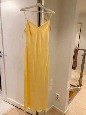 Ljusgul klänning  - Säljer en ljusgul långklänning med smala axelband och v-ringad hals. Klänningen har en enkel, rak passform och är tillverkad i ett glansigt material som ger en lyxig känsla. Perfekt för sommarens alla tillfällen! Aldrig använd 