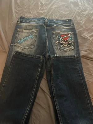 Ed Hardy jeans med brodyr och tryck - Säljer ett par blå jeans från Ed Hardy med coolt dödskalletryck och röd hatt på ena bakfickan samt broderad logga på den andra. Jeansen har raka ben, slitningar och kontrastsömmar. Perfekta för dig som gillar streetwear och detaljer. Dem sitter bra.