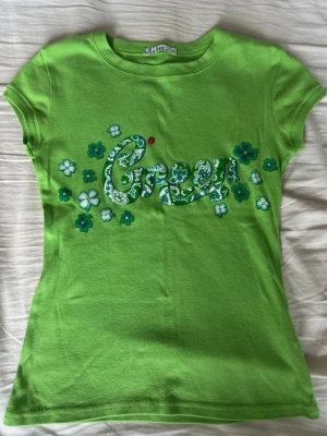 Grön y2k topp - Grön y2k topp med jättefina detaljer på framsidan. Observera fläcken vid texten (har ej lyckats få bort den) samt tyget åkt upp som en tråd på baksidan (se bilder) dock inget direkt märkbart när man har toppen på sig.