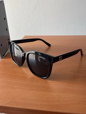 Svarta solglasögon från Gucci - Stilrena svarta solglasögon från Gucci med fyrkantig form och breda skalmar. Klassisk design med diskret logga på sidan och mörka glas. Perfekta för att lyfta din look med en lyxig touch.