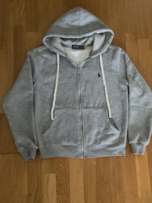Grå zip hoodie från Polo Ralph Lauren - Snygg grå hoodie med dragkedja från Polo Ralph Lauren. Klassisk design med vit dragsko, två fickor framtill och broderad logga på bröstet. Mjuk insida i fleece och ribbade muddar vid ärmar och nederkant. Perfekt för chill dagar.