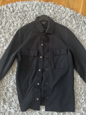 Svart overshirt med tryckknappar - Stilren svart overshirt med klassisk krage och stora bröstfickor med lock. Jackan har tryckknappar framtill och är gjord i ett tjockare tyg som ger en schysst struktur. Perfekt lager-på-lager-plagg för en avslappnad look. Den är aningens nopprig men går säkert att få bort med en noppborttagare.