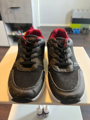 Svarta Kappa sneakers med röd detalj - Snygga svarta sneakers från Kappa med röda detaljer och vit sula. Skorna har mesh- och syntetmaterial på ovandelen, klassisk snörning och låg profil. Perfekta för en sportig och avslappnad stil.