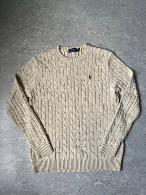 Beige kabelstickad tröja Polo Ralph Lauren - Kabelstickad tröja i beige från Polo Ralph Lauren med rund halsringning och klassisk broderad logga på bröstet. Tröjan har långa ärmar och ribbade muddar vid ärmslut och nederkant. Perfekt för en stilren och avslappnad look.
