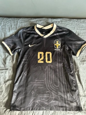 Svart Brasilien matchtröja Nike - Snygg svart Brasilien matchtröja från Nike med guldiga detaljer och nummer 20 på både fram- och baksidan. Tröjan har korta ärmar, V-ringad krage med vita kanter och Brasilien-emblem på bröstet. Perfekt för fotboll eller streetwear.