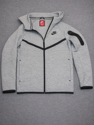 Grå Nike hoodie med dragkedja - Snygg grå hoodie från Nike med hel dragkedja framtill och svarta detaljer. Har två sidofickor med dragkedja och en extra ficka på ärmen. Nike-logga på bröstet och sportig design. Perfekt för en avslappnad streetwear-look.