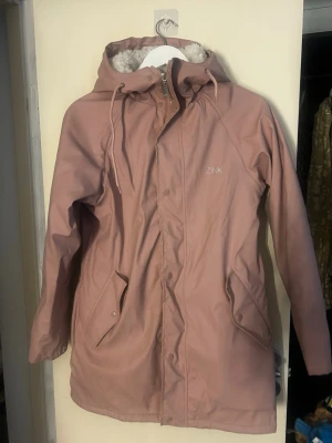 Rosa regnjacka med teddyfoder från Zink - Snygg rosa regnjacka från Zink med mysigt vitt teddyfoder (inte foder i armarna). Jackan har huva med dragsko, två framfickor med lock och stängs med dragkedja och tryckknappar. Perfekt för regniga dagar när du vill hålla dig både torr och varm. Använd 2 gånger