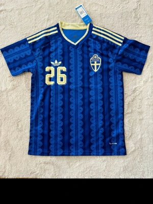 Blå Sverige fotbollströja Adidas - Säljer en blå fotbollströja från Adidas med Sveriges landslagsemblem och nummer 26 i gult. Tröjan har gula detaljer på axlarna, mönstrad design och namnet 'Ali' på ryggen. Tillverkad i lätt och ventilerande material, perfekt för träning eller match.