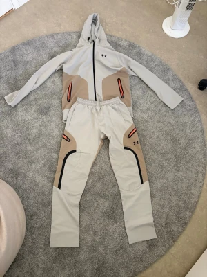 Beige och grått loungewear-set Under Armour - Snyggt loungewear-set från Under Armour i beige och grått med svarta och orange detaljer. Setet består av en hoodie med dragkedja och matchande byxor med resår i midjan. Båda plaggen har coola kontrastpaneler och logga på bröst och ben. Perfekt för chill dagar hemma. Kan säljas separat! 
