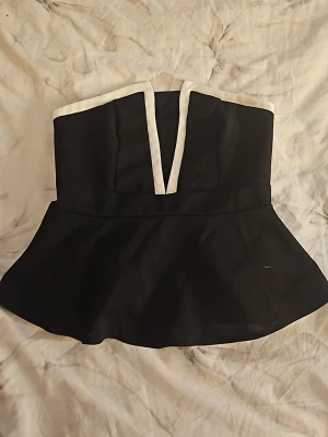 Svart peplum tubtopp med vit kant - Snygg svart peplum tubtopp med vit kant längs överdelen och ett V-format snitt framtill. Toppen har en figurnära passform upptill och volang nertill. Smockad rygg för extra komfort. Perfekt för att skapa en trendig look.