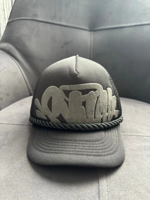 Svart truckerkeps med graffiti-tryck - Snygg svart truckerkeps med stort graffiti-inspirerat tryck framtill och broderad text på skärmen. Kepsen har mesh på sidorna och bak, samt ett repdetalj ovanför skärmen för extra streetwear-vibe. Justerbar baktill för perfekt passform.