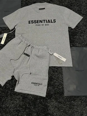 (Separat)Grått Essentials set med t-shirt & shorts - Säljer ett grått set från Essentials Fear of God med t-shirt och matchande shorts. Båda plaggen har svart logotyptryck framtill och är tillverkade i mjuk bomull för en avslappnad och bekväm känsla. Perfekt för en chill och stilren look.