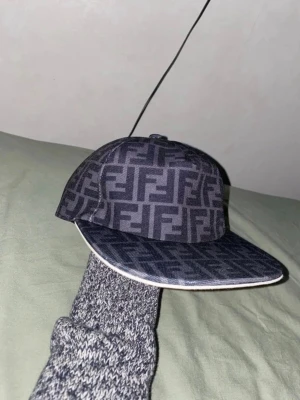 Svart Fendi keps - Snygg svart keps med tydligt FF-mönster över hela ytan. Klassisk böjd skärm och justerbar rem baktill. Kepsen har en modern streetwear-vibe och är tillverkad i ett slitstarkt tygmaterial.