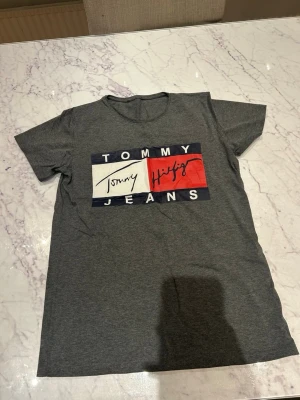 Grå Tommy Jeans t-shirt - Snygg grå t-shirt från Tommy Jeans med klassisk logga framtill i vitt, rött och marinblått. T-shirten har rund halsringning och korta ärmar. Perfekt till en avslappnad stil och enkel att matcha med jeans eller shorts.