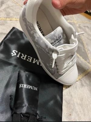 Vita och grå sneakers från Numeris - Snygga sneakers från Numeris i vitt och grått med digitalt camomönster på sidorna. Skorna har vita snören, vit tå och sula samt en låg profil. Tillverkade i textil och syntetmaterial. Perfekta för dig som gillar en clean och modern look.
