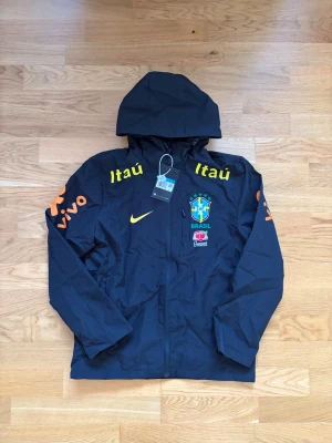Svart Brasil Nike vindjacka med huva - Snygg svart vindjacka från Nike med Brasil-logga och sponsormärken i gult och orange. Jackan har huva, dragkedja framtill och tryck på bröst och ärmar. Perfekt för dig som gillar fotboll och sportig stil.