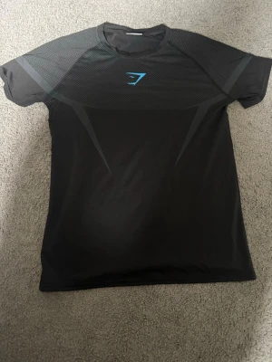 Svart träningströja från Gymshark - Snygg svart träningströja från Gymshark med blå logga på bröstet. Tröjan har korta ärmar och meshdetaljer upptill för extra ventilation. Tillverkad i ett lätt och stretchigt funktionsmaterial som passar perfekt till gymmet eller löprundan.