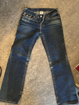 True Religion blå jeans med kontrastsömmar - Säljer ett par klassiska vintage blå jeans från True Religion med tydliga kontrastsömmar och ikoniska bakfickor med vita knappar. Jeansen har normal passform och bootcut. Materialet är slitstarkt denim i bomull och de har en snygg tvättad look. Köpta för 2100kr. 