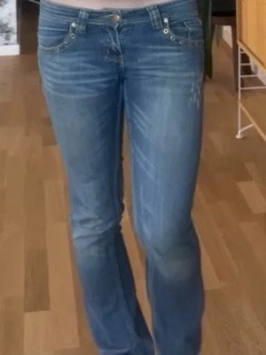 Blå bootcut jeans med broderade fickor - Snygga blå bootcut jeans med slitningar och detaljerade broderier i guld på bakfickorna. Jeansen har låg midja, klassisk femficksmodell och stängs med dragkedja och knapp. Köpta på fransk loppis men passar tyvärr inte längre.                Ena bakfickan saknar knapp!!