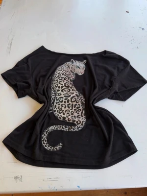 Svart t-shirt med leopardtryck - Cool svart offshoulder t-shirt med korta ärmar och ett stort leopardtryck på ryggen. T-shirten har en loose passform och är gjord i mjuk bomull. Perfekt för dig som vill sticka ut med ett djuriskt motiv.