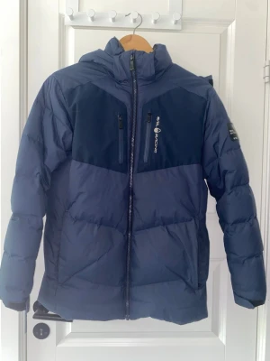 Mörkblå dunjacka Sail Racing - Snygg mörkblå dunjacka från Sail Racing med två bröstfickor med dragkedja, hög krage och huva. Jackan har en sportig look med logga på bröstet och ärmen. Perfekt för kalla vinterdagar tack vare det tjocka dunfodret. Pris kan diskuteras.