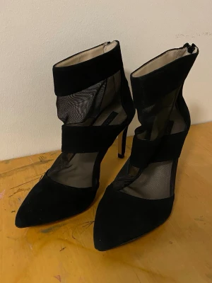 Svarta mesh pumps med spetsig tå - Säljer ett par svarta pumps med spetsig tå och höga smala klackar. Skorna har coola meshdetaljer och breda sammetsband runt foten och ankeln. Dragkedja baktill för enkel påtagning. Perfekta för dig som vill sticka ut med en edgy look.