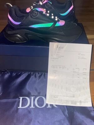 Svarta Dior sneakers med neon-detaljer - Chunky svarta Dior sneakers med mesh och läder, samt neonfärgade detaljer i lila, rosa och grönt. Snygg design med robust sula och CD-logga undertill. Skorna har metalliska öglor och reflekterande partier som ger en futuristisk vibe.