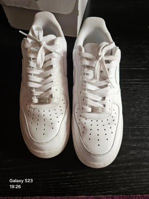 Vita Nike Air Force 1 sneakers - Klassiska vita Nike Air Force 1 sneakers i lågt utförande. Skorna har en stilren design med perforerad tå, kraftig sula och ikoniska Swoosh-loggan på sidan. Tillverkade i läder med textildetaljer och vita skosnören. Perfekta för en clean och tidlös look.