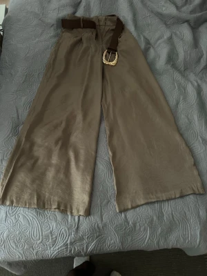 Beiga vida linnebyxor från Zara med bälte - Säljer brun/beiga byxor från Zara med breda ben och hög midja. Byxorna har ett brunt bälte med guldfärgat spänne som detalj. Perfekta för en avslappnad och trendig look. Materialet är svalt och har en snygg lyster.
