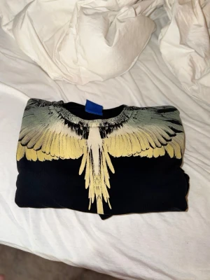 Marcelo burlon sweatshirt - Säljer en svart topp med ett stort, detaljerat vingmönster i gult och grått över axlar och bröst. Tröjan har rund halsringning och lång ärm. Perfekt för dig som vill sticka ut med en unik design.