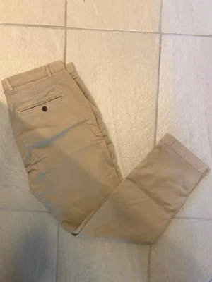 Beige chinos Morris herr  - Snygga beige chinos med rak passform och broderad detalj ovanför bakfickan. Byxorna har bälteshällor, knappstängning på bakfickan och är tillverkade i ett mjukt bomullsmaterial. Perfekta för en clean och stilren look.