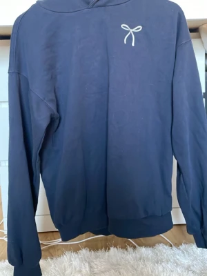 Mörkblå hoodie med rosetttryck - Snygg mörkblå hoodie med vit rosetttryck på bröstet och ett större rosett- och hjärtmotiv på ryggen. Hoodien har huva och ribbade muddar vid ärmslut och nederkant. Perfekt för en chill och trendig look.