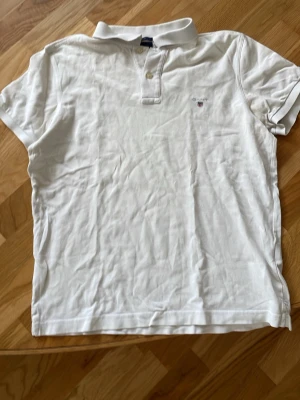Vit piké från Gant i bomull - Klassisk vit pikétröja från Gant i bomull med korta ärmar och liten logga broderad på bröstet. Regular fit och tidlös design med krage och knappar framtill. Perfekt till jeans eller shorts för en clean look.