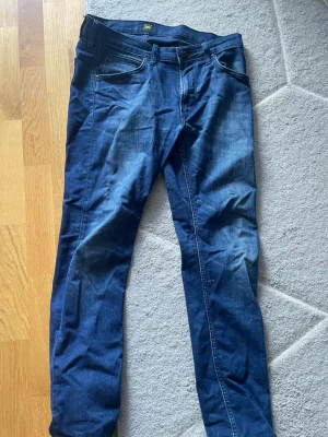 Mörkblå jeans från Lee - Säljer ett par mörkblå jeans från Lee med klassisk femficksdesign och raka ben. Jeansen har snygga kontrastsömmar och är tillverkade i ett mjukt denimtyg som sitter skönt. Perfekta för dig som gillar en tidlös och clean look.