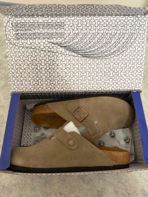 Taupe Birkenstock Boston 38 - Säljer ett par klassiska Birkenstock Boston tofflor i beige mocka med justerbart spänne i metall. Tofflorna har en bekväm korksula och mjuk fotbädd som formar sig efter foten. Perfekta för dig som gillar stilren och avslappnad look. ‼️Skickar inom 24H‼️ **PRIS KAN DISKUTERAS**