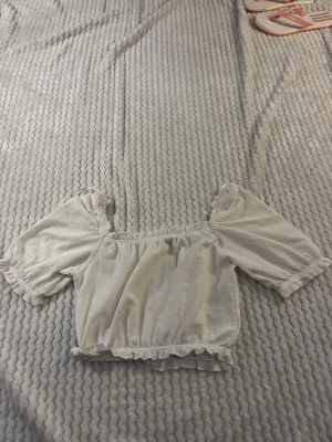 Vit croppad blus med volang - Söt vit croppad blus med volangdetaljer och puffiga korta ärmar. Toppen har resår vid axlar och midja för en bekväm passform och en romantisk look. Perfekt att styla med jeans eller kjol för en trendig outfit.