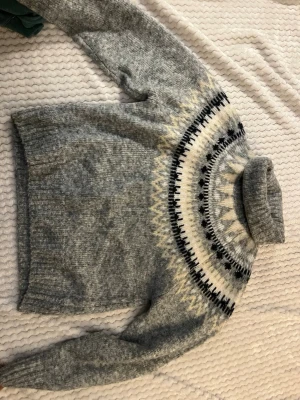 Grå polotröja med nordiskt mönster - Mysig grå stickad polotröja med klassiskt nordiskt mönster i vitt, svart och beige runt halsen. Tröjan har lång ärm och ribbstickade muddar. Perfekt för kalla dagar och ger en stilren vinterlook.