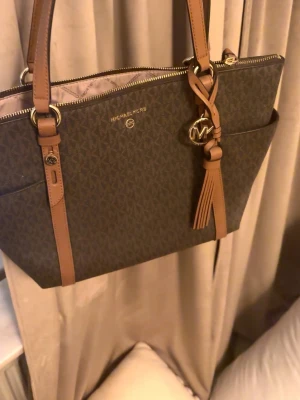 Brun handväska från Michael Kors - Snygg brun handväska från Michael Kors med klassiskt MK-mönster, ljusbruna detaljer och axelremmar. Väskan har guldfärgade metalldetaljer, tofs och MK-hänge. Perfekt för dig som vill ha en trendig och rymlig väska i tåligt material.