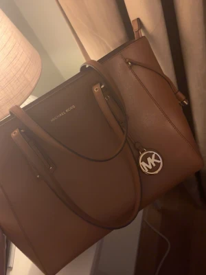 Brun toteväska från Michael Kors - Snygg brun handväska från Michael Kors i klassisk tote-modell. Väskan har guldfärgade detaljer, dubbla handtag och en rund MK-berlock. Tillverkad i tåligt läder med dragkedja upptill och flera innerfack. Perfekt för dig som vill ha plats för allt du behöver.
