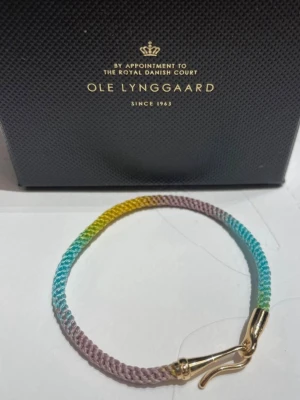 Flerfärgat armband Ole Lynggaard - Snyggt armband från Ole Lynggaard i flätad textil med färgskiftningar i lila, blått, grönt och gult. Storlek 16 cm . Spännet är i 18 karats guld och har en elegant krokdesign. Perfekt för dig som gillar färg och exklusiva detaljer. Nypris 9200 . Armbandet är ej använt och kommer i original box samt påse. 