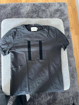 Svart t-shirt från Les Deux - Säljer en svart t-shirt från Les Deux med två vertikala sammetsränder på bröstet. Klassisk rund halsringning och korta ärmar. Perfekt för dig som gillar stilrena plagg med en twist.
